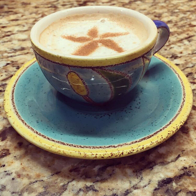 A colorful ceramic cup filled with a creamy beverage, featuring a decorative star pattern on the frothy surface, sits on a matching saucer against a textured granite countertop.